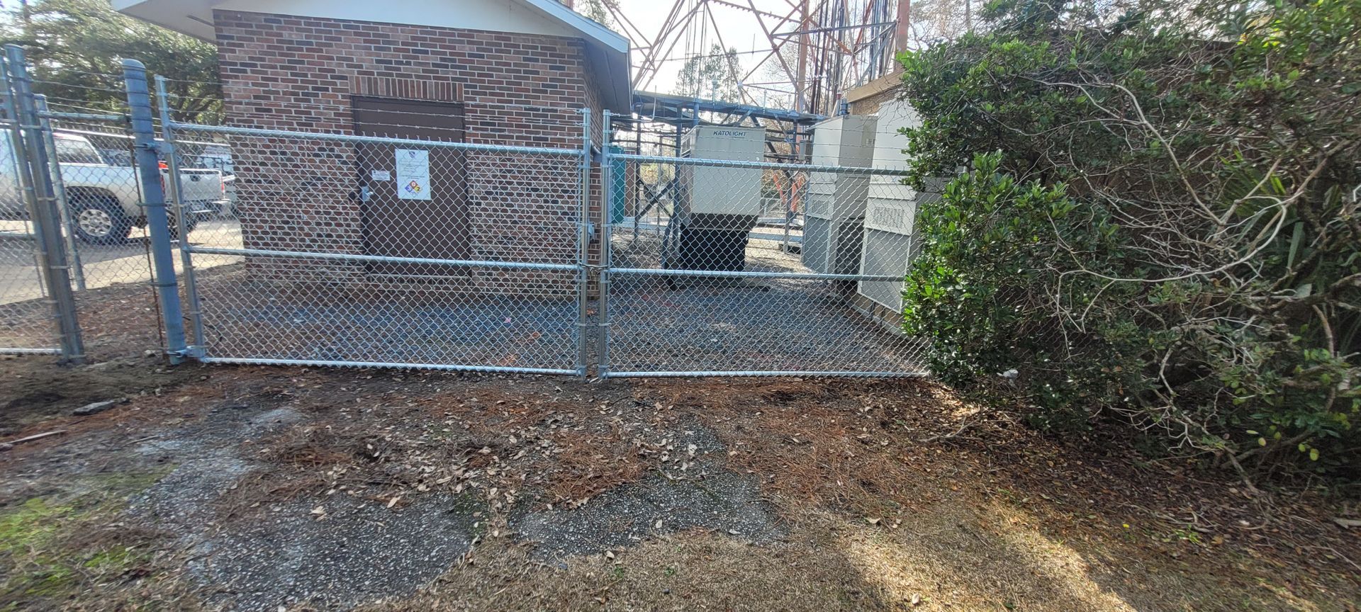 A chain-link fence surrounds a utility building. A pickup truck is parked nearby. Green shrubbery is to the right.