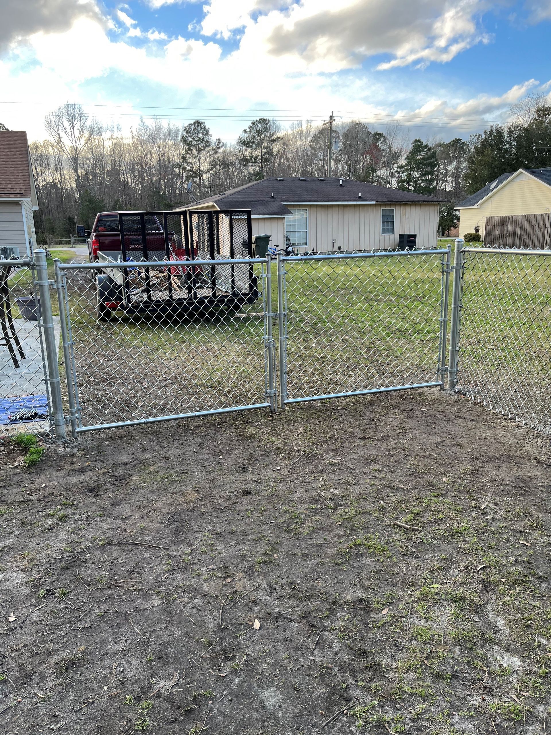Chain-link fence in front of a house. A lawn is in the background with a trailer. Cloudy sky.