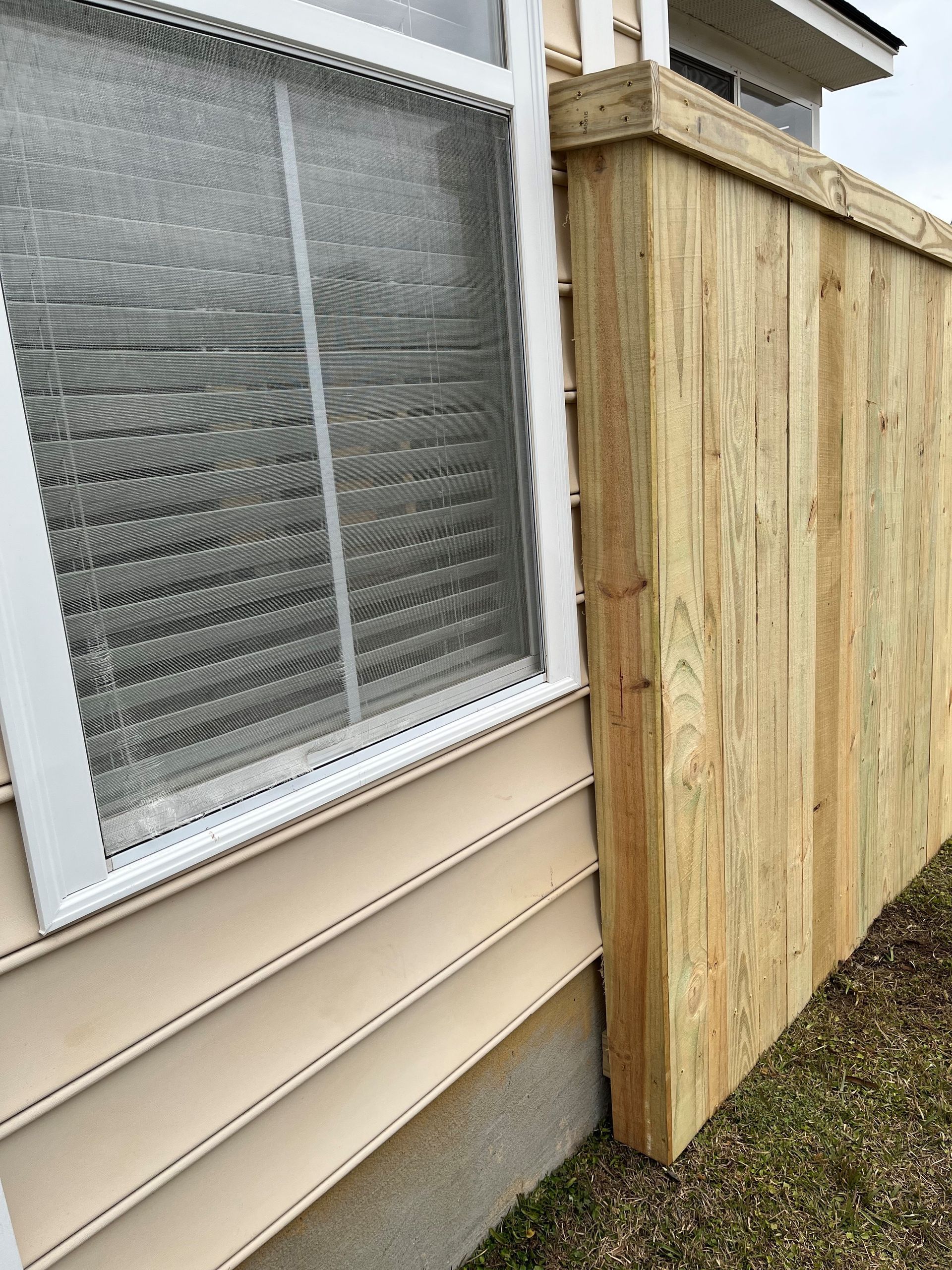 Wooden fence built next to a building's window, beige siding, and green grass.