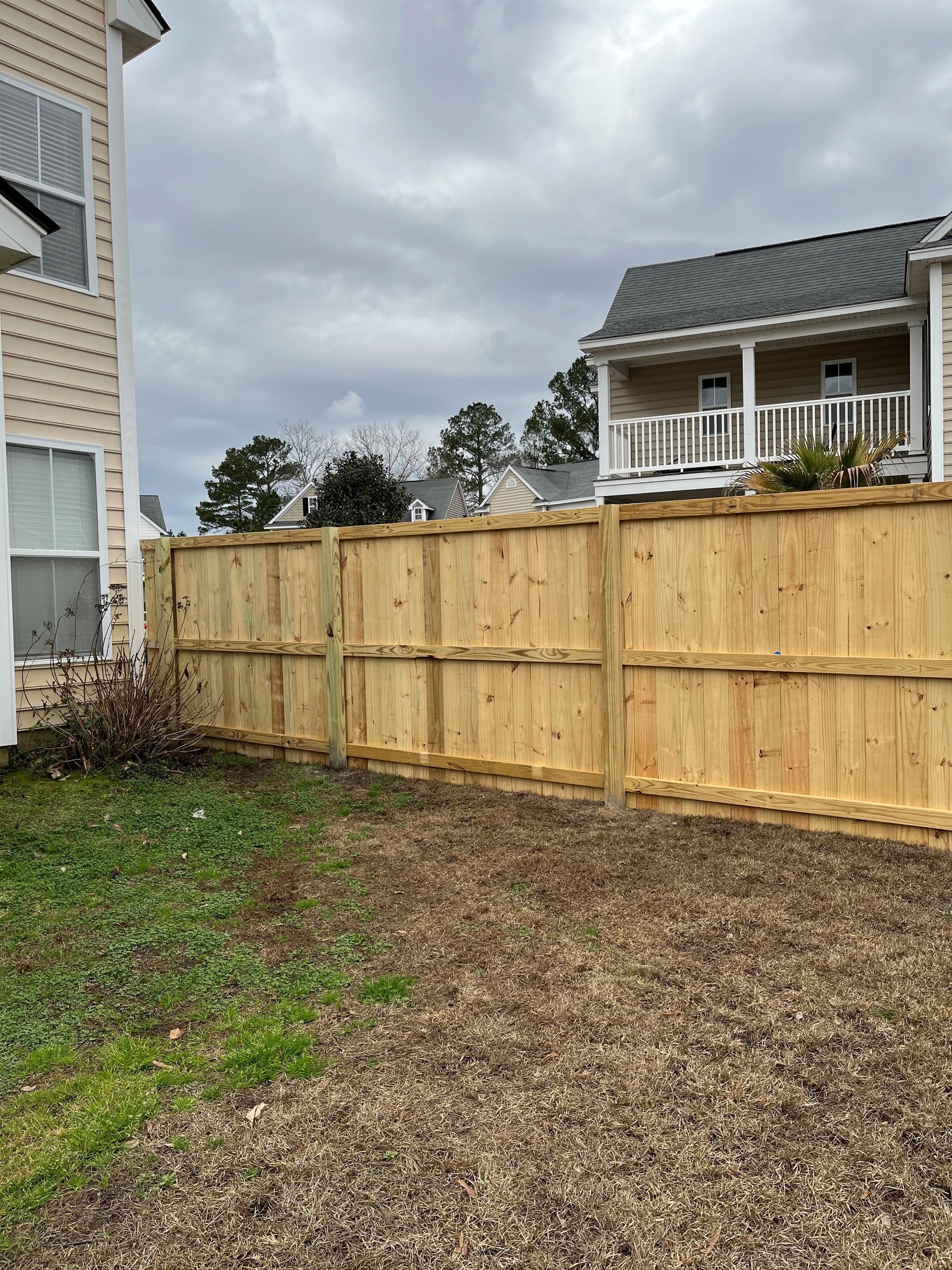 Wooden fence separating two beige-colored houses, with a yard of dirt and grass in front. Cloudy sky.