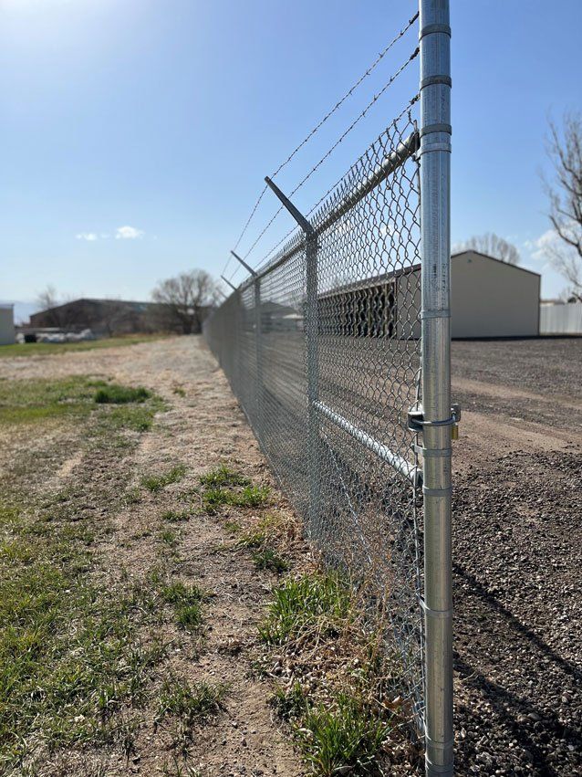 Chain-link fence with barbed wire on top, bordering a gravel road and grassy area under a sunny sky.
