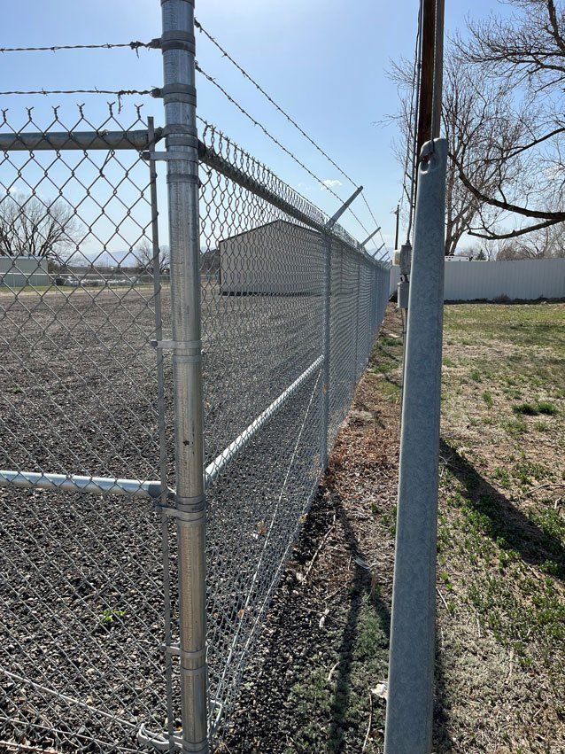 Chain link fence with barbed wire on top, enclosing a gravel area, under a bright sky.