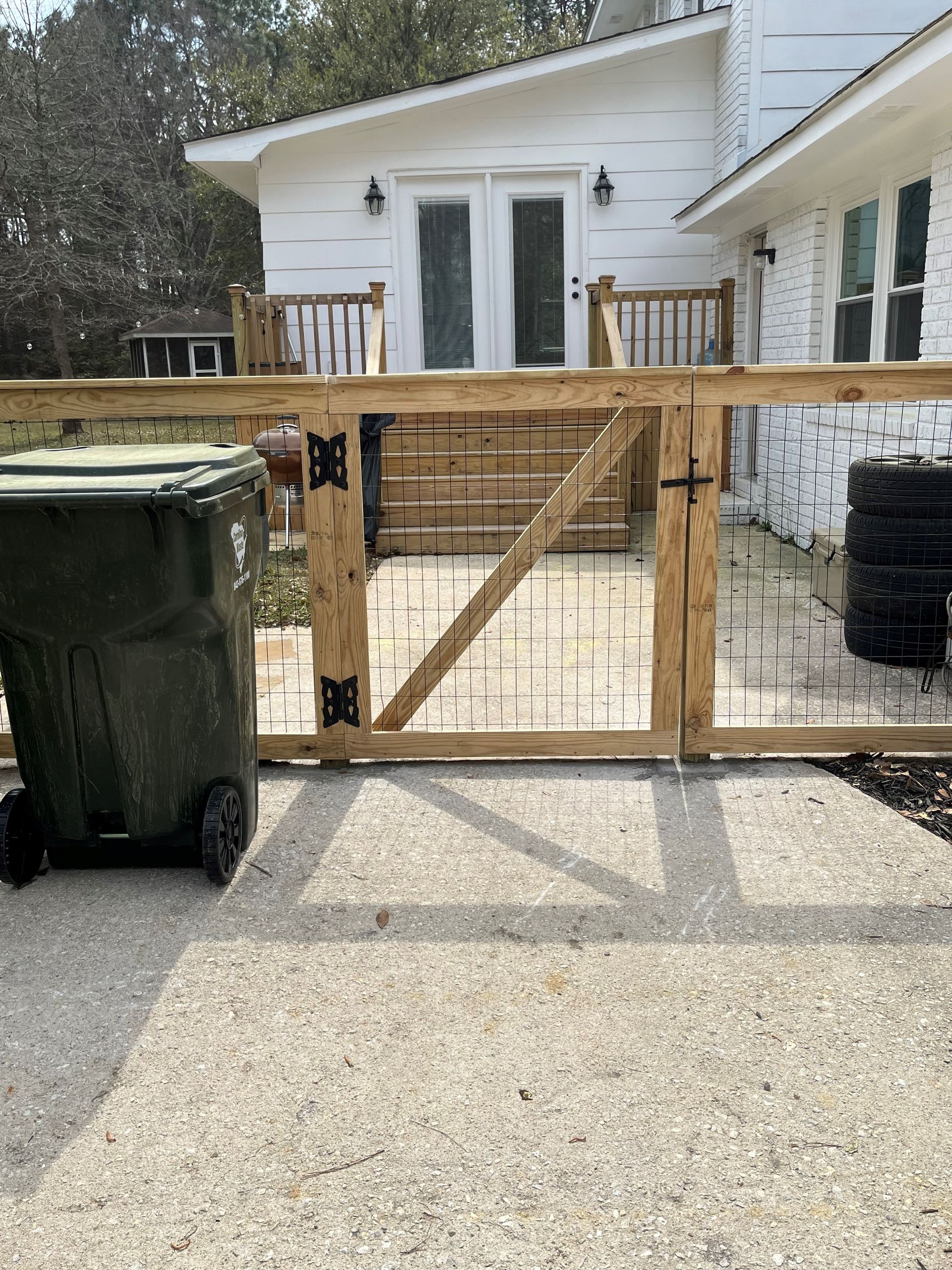 Wooden gate and fence with a trash can in front of a white house with a deck.