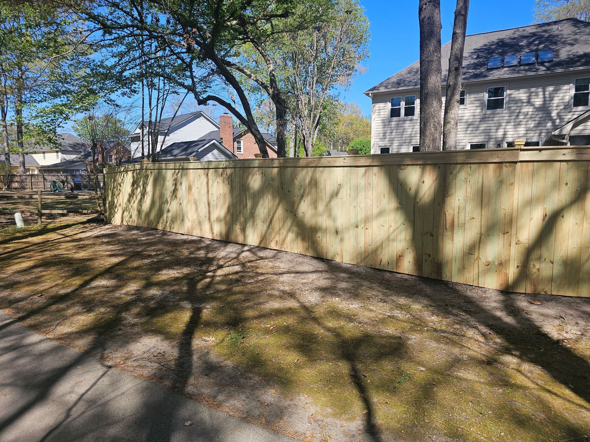 Wooden fence in yard, with shadows cast across grass and asphalt. Homes in background.