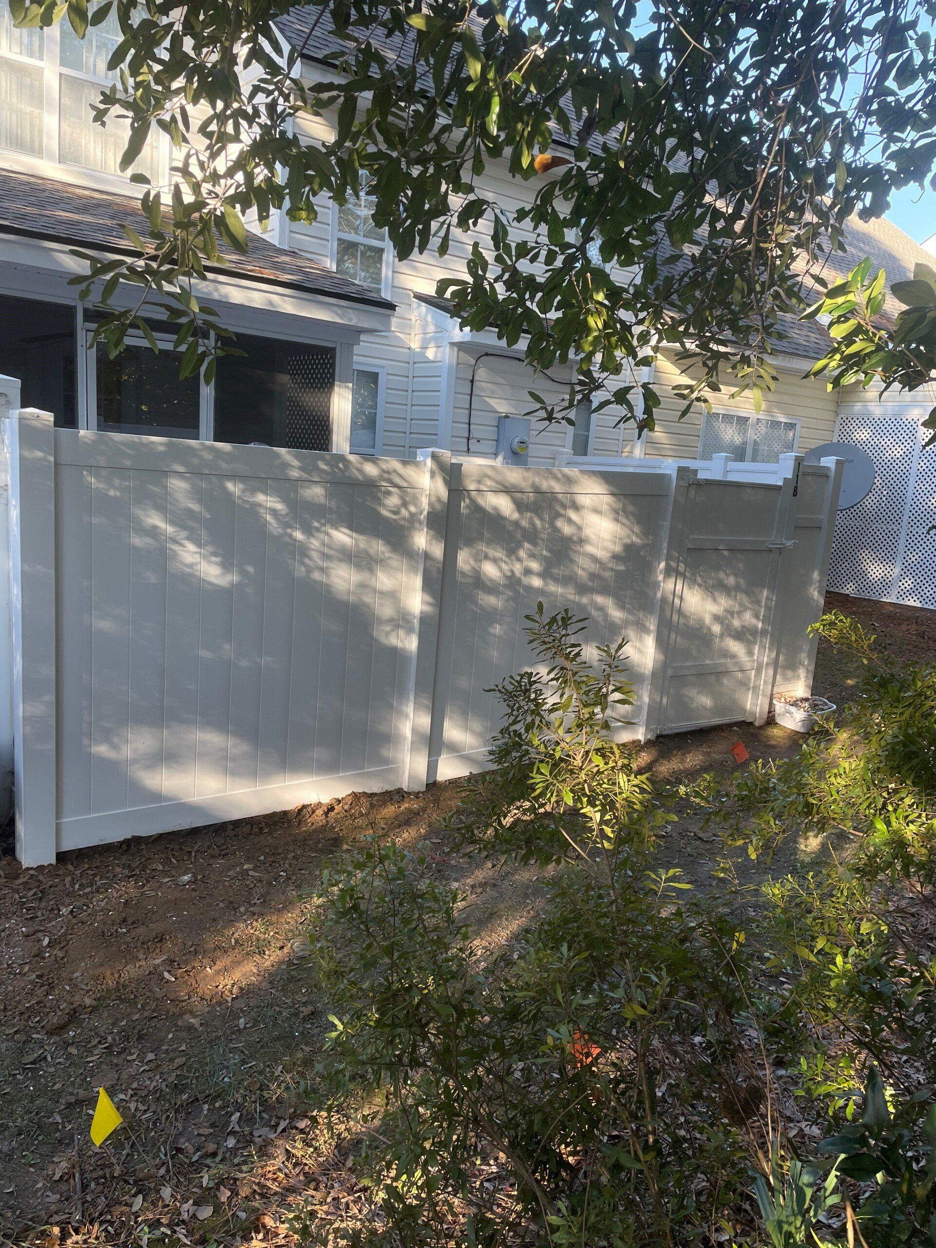 Beige vinyl fence near a house, with foliage in front and above. Sunny outdoors.