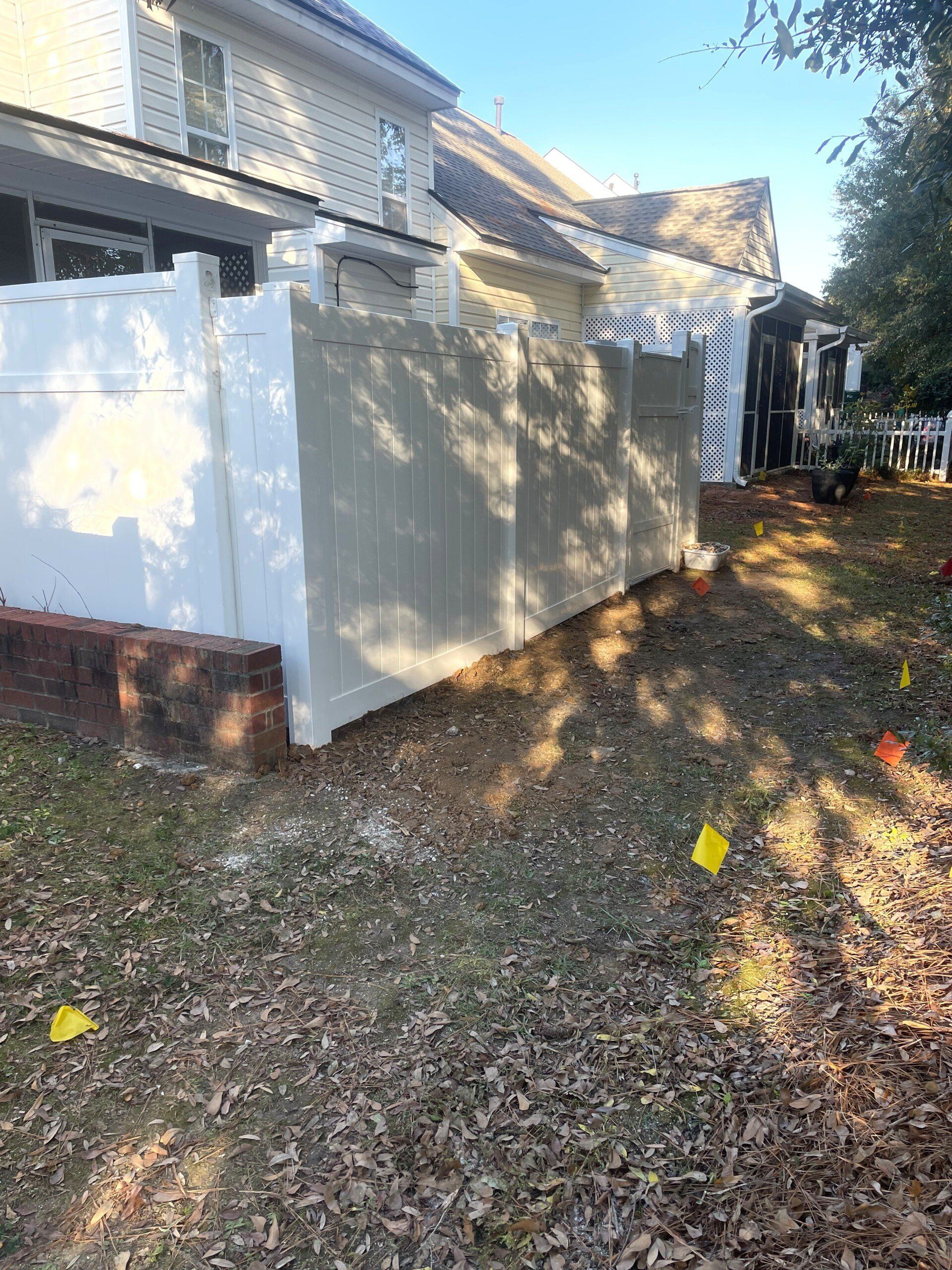 White fence bordering a yard next to a beige house, with fall foliage and yellow flags.