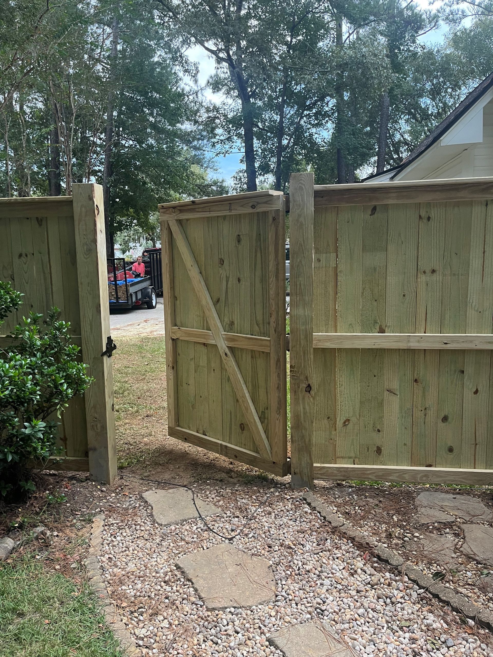 Wooden gate, partially open, within a wooden fence, leading to a yard with a vehicle.