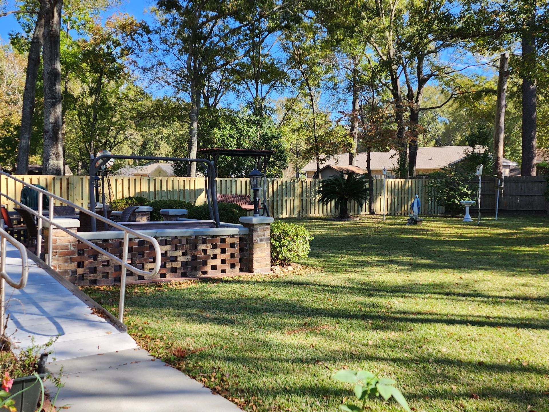 A sunny outdoor area with a brick structure, fence, and trees. Grassy area, walkway, and sky.