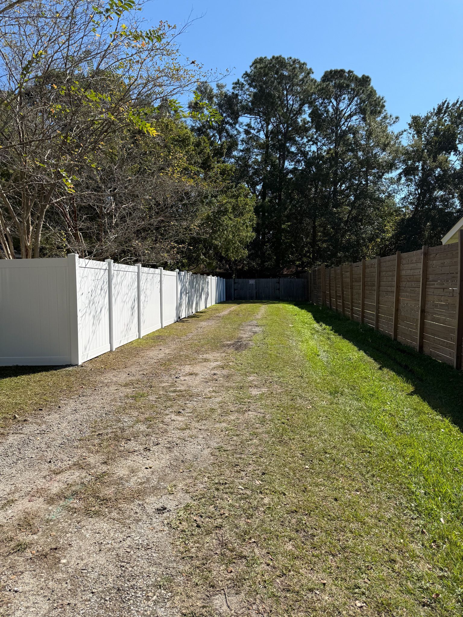 Gravel driveway between two fences. White fence on the left, brown fence on the right, trees in the background.