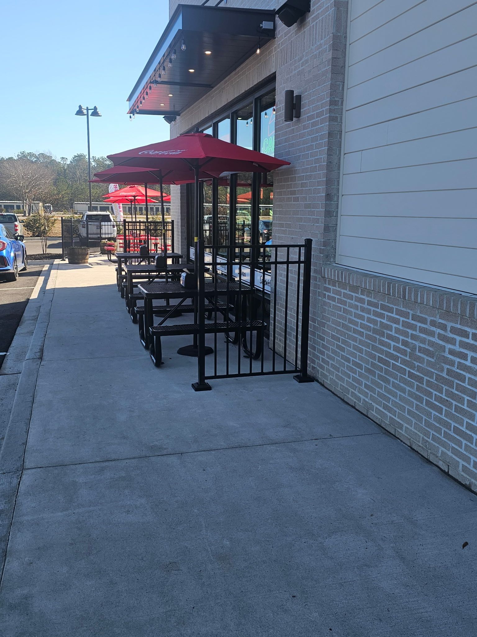 Outdoor seating area with tables, red umbrellas, and black metal fences. Adjacent to a building with brick facade.