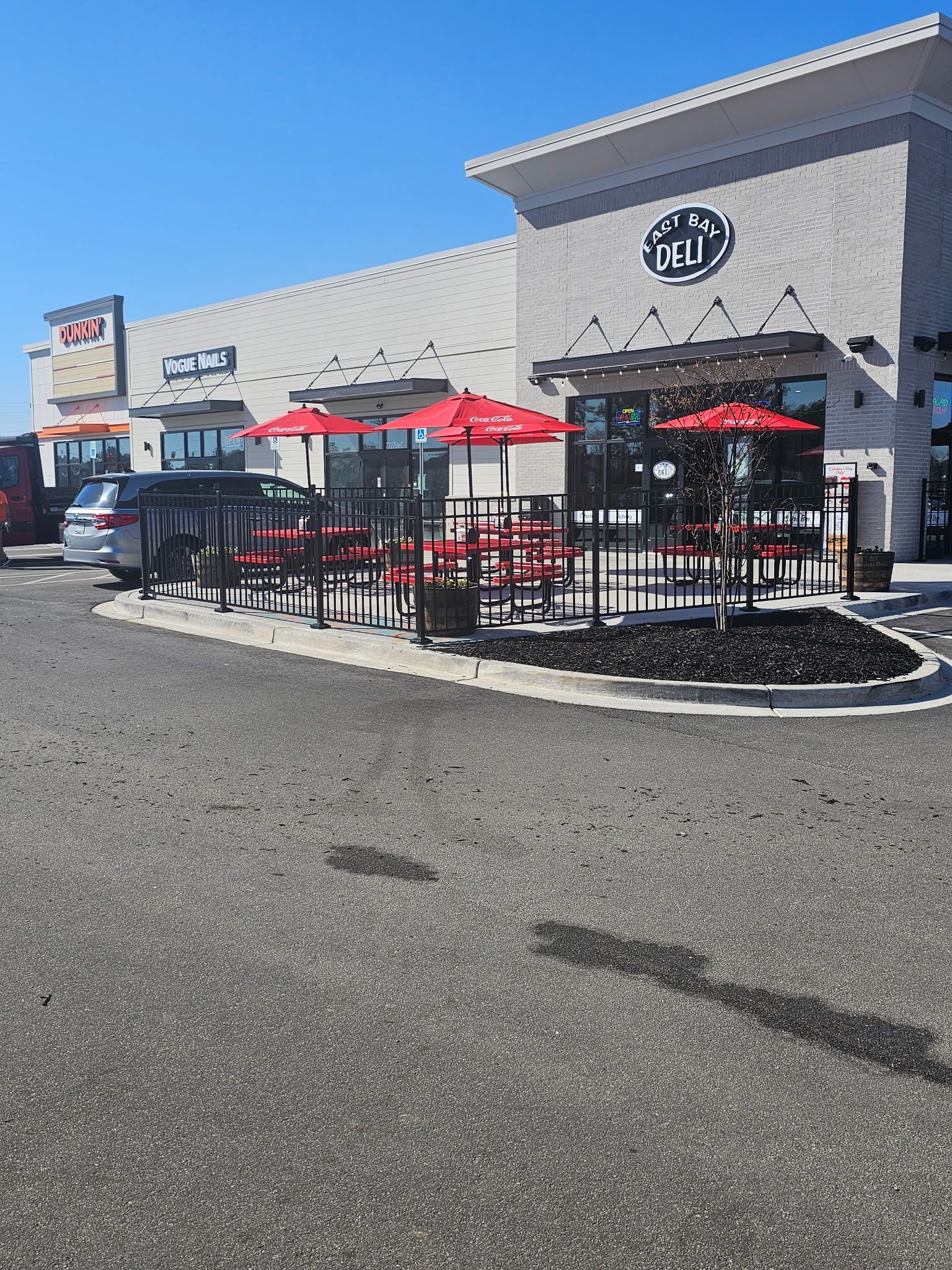 Restaurant exterior with outdoor seating: red umbrellas, black fences, and a bright blue sky.