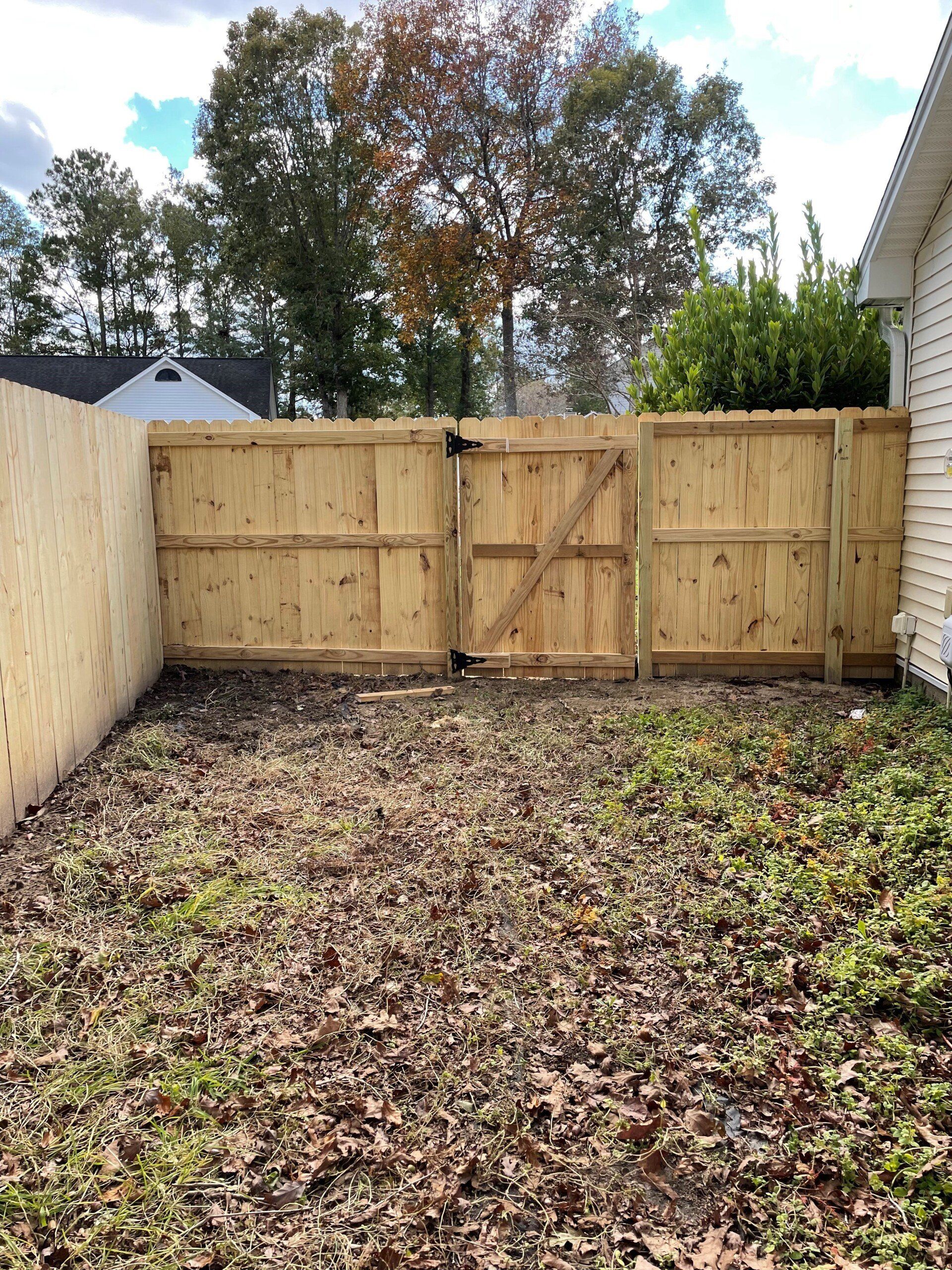 Wooden gate in a backyard enclosed by a wooden fence, with dried leaves and low ground cover.