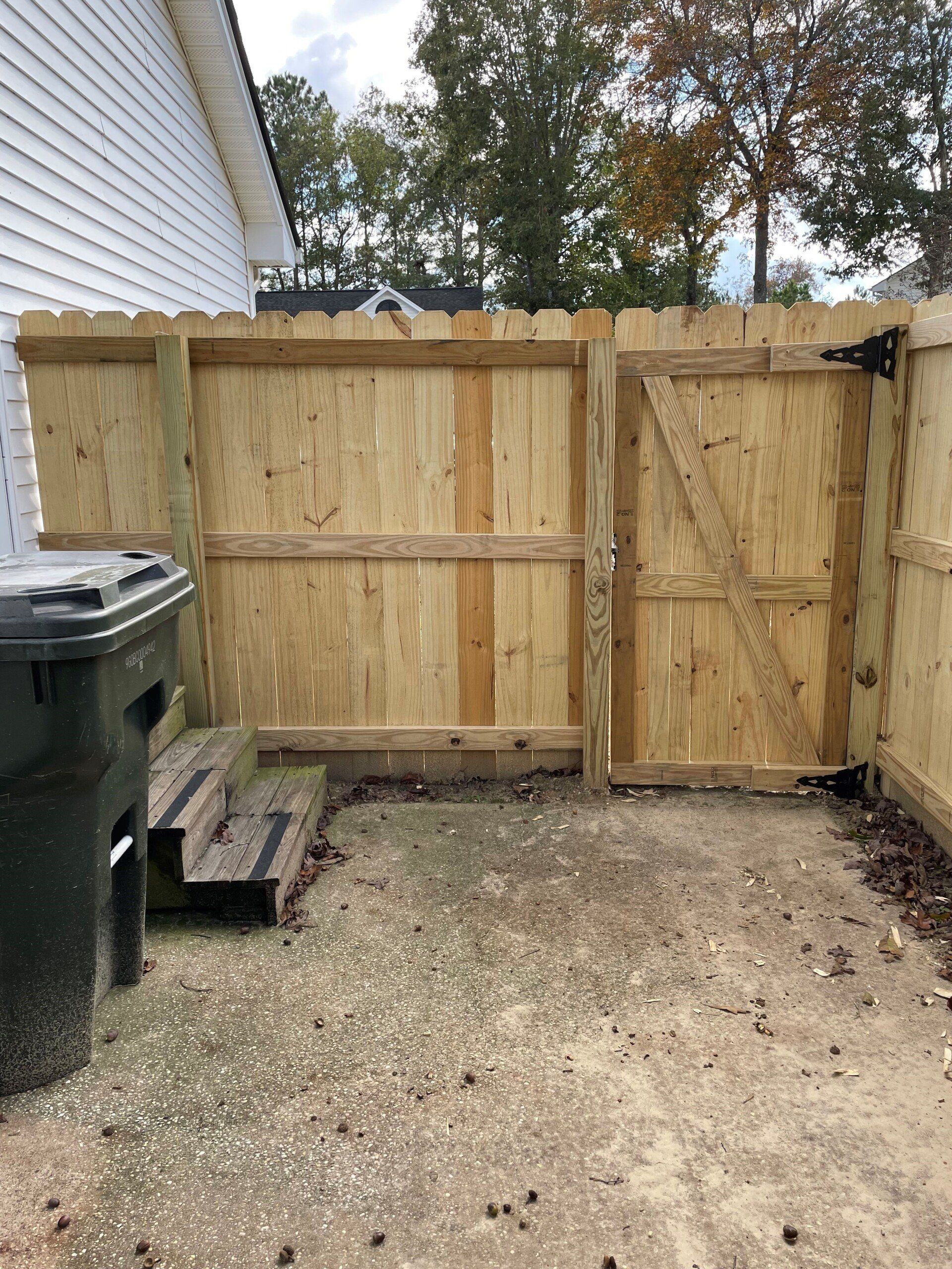 Wooden backyard fence with gate. Steps lead to a small concrete area; trash can on the left.