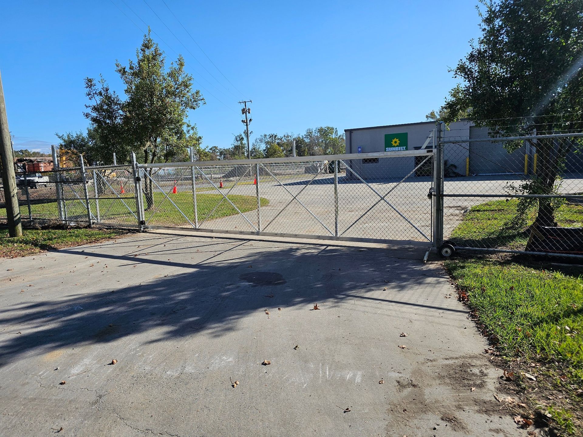 A chain-link fence with a closed gate leading to a paved area and a building under a blue sky.