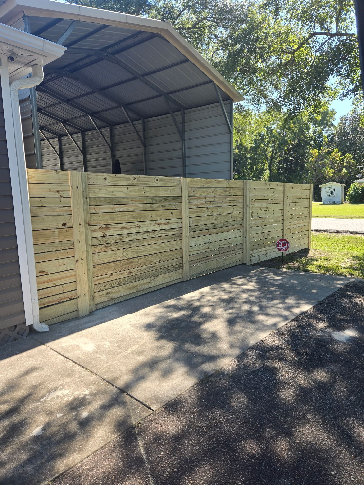 A wooden fence with a carport in the background on a sunny day.