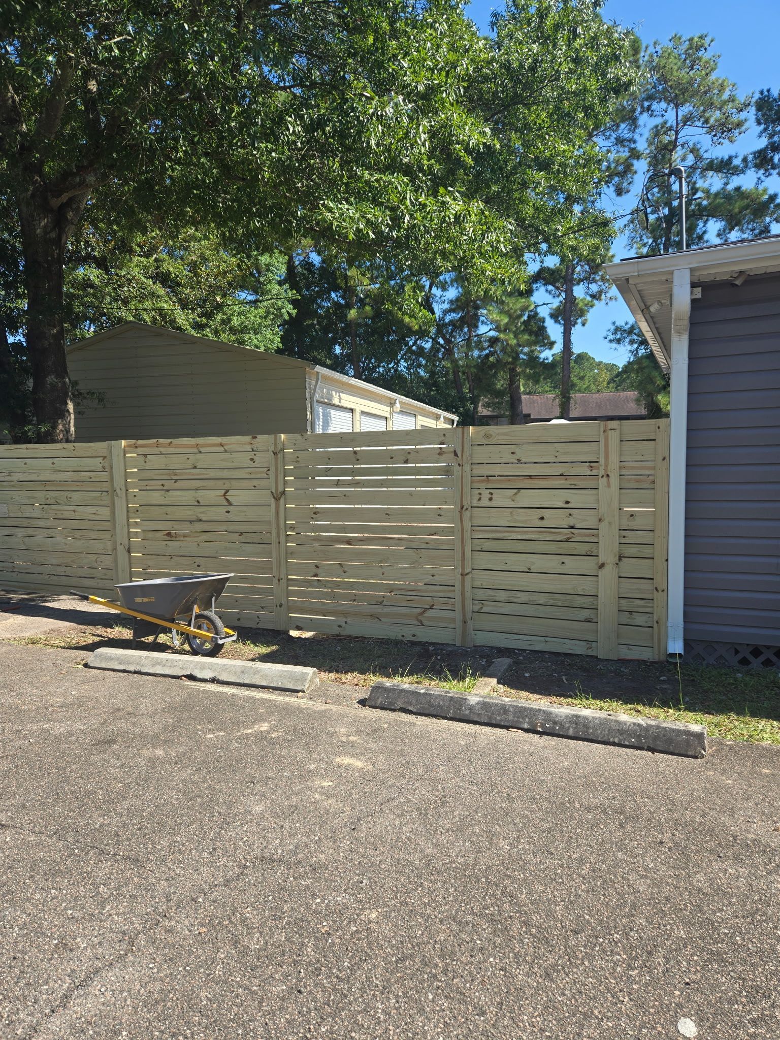 A wooden fence with horizontal slats in front of a building. A wheelbarrow sits nearby on the asphalt.