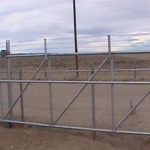 Metal chain-link fence topped with barbed wire, outdoors. Brown earth and cloudy sky in the background.