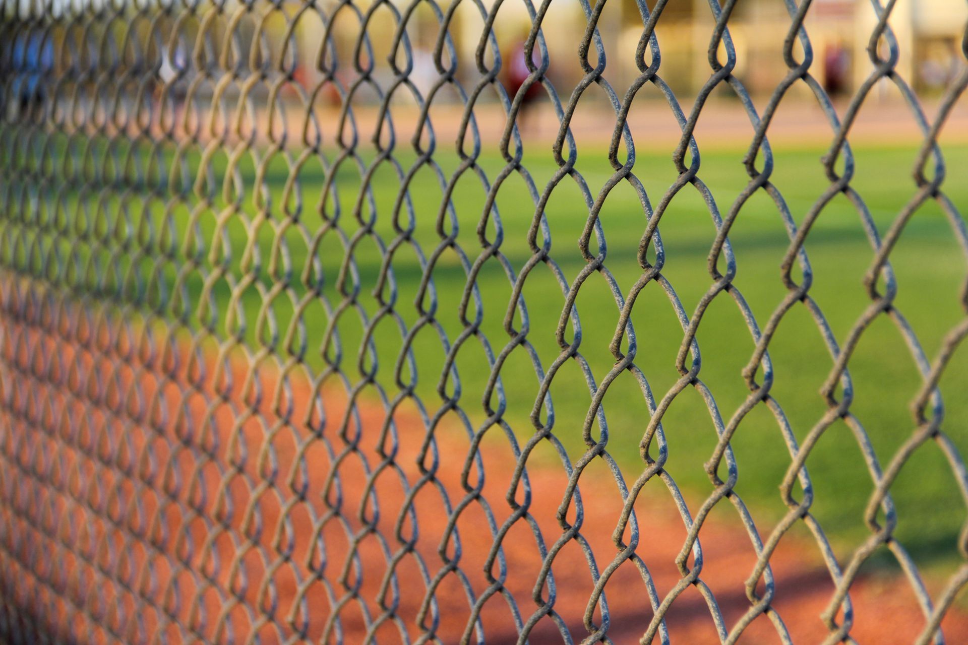 Chain-link fence in foreground, baseball field blurred in background; green grass, brown dirt.