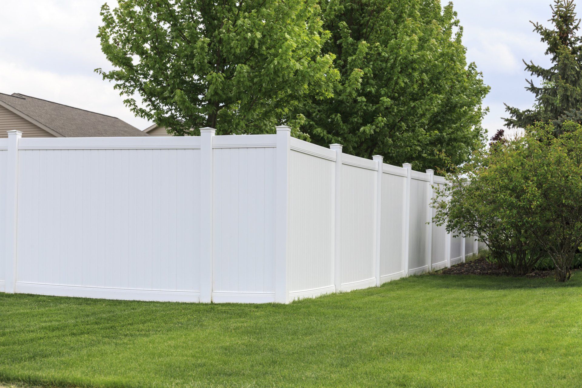 White vinyl fence surrounding a grassy lawn with trees in the background under a cloudy sky.