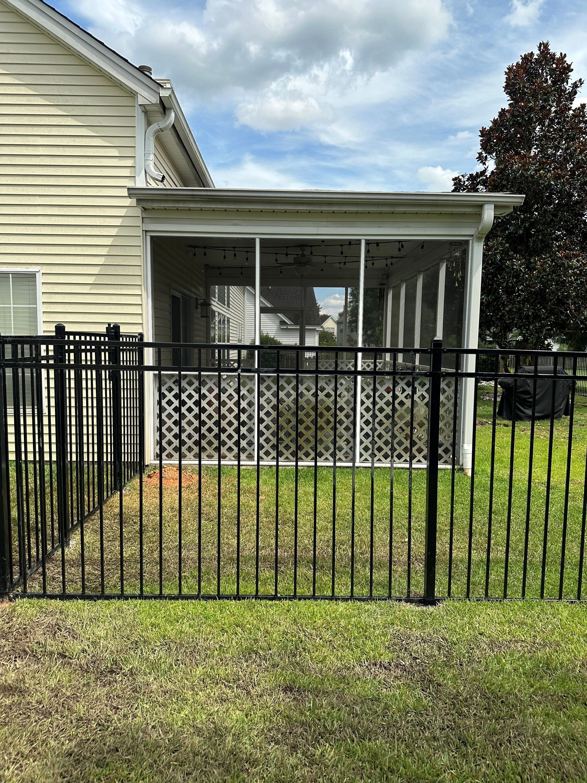 Black fence surrounds a screened porch attached to a beige house, with green grass and cloudy sky.