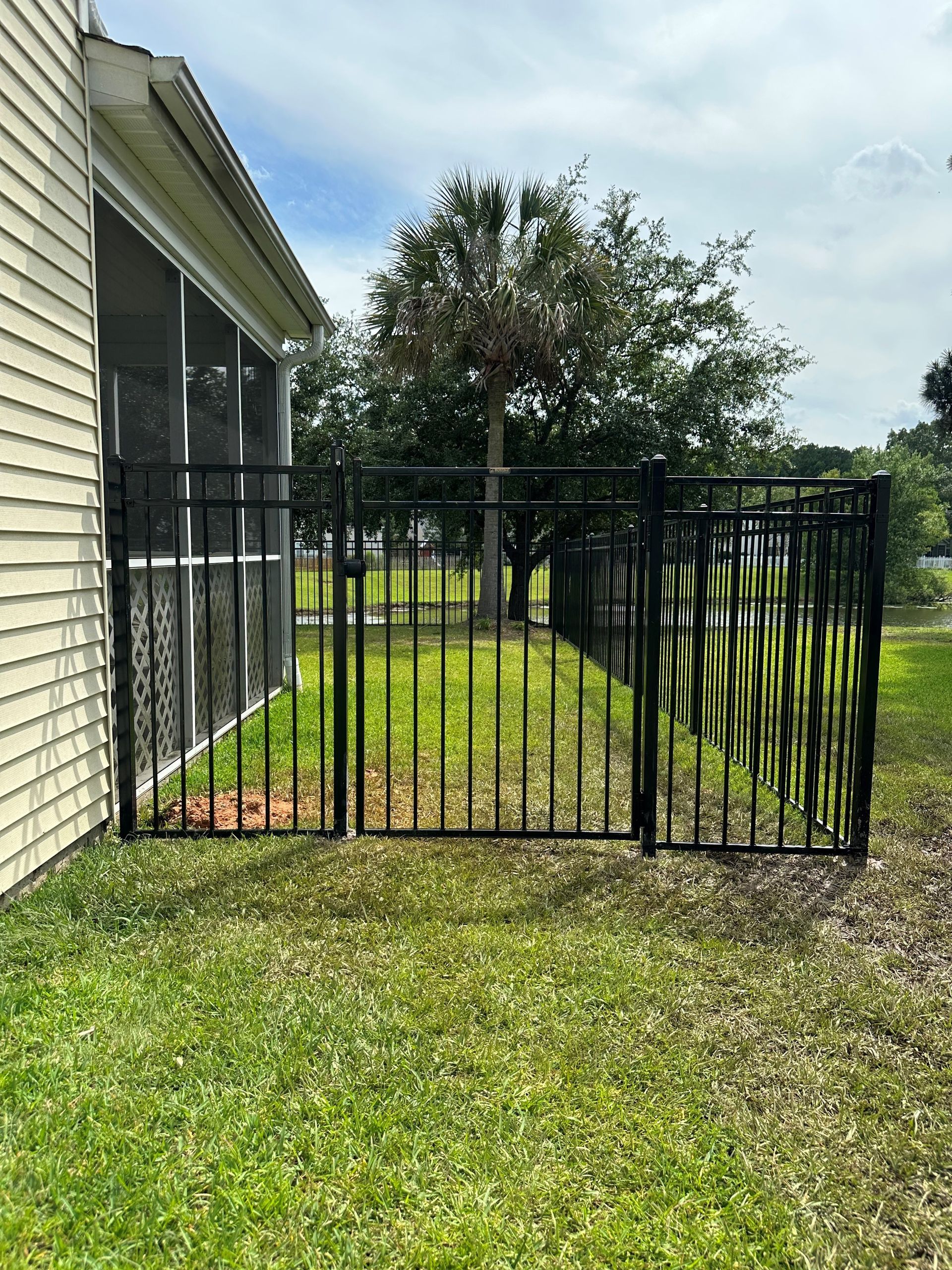 Black metal fence encloses a section of yard next to a beige house, green grass, and a tree.