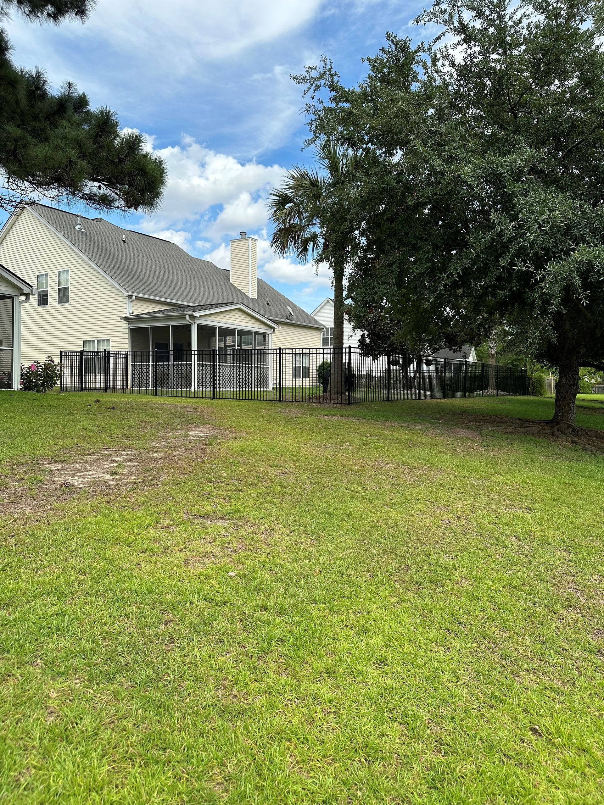 Lawn in front of a light-colored house with a black fence, trees, and a partly cloudy sky.