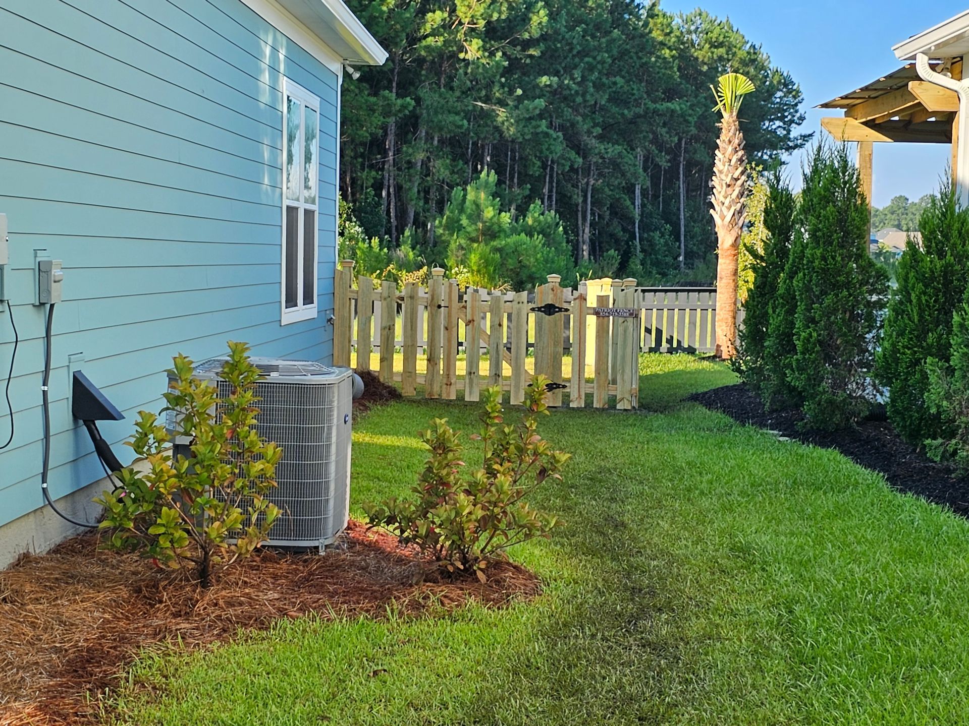 Side yard of a house with light blue siding, air conditioning unit, wooden fence, and green grass.