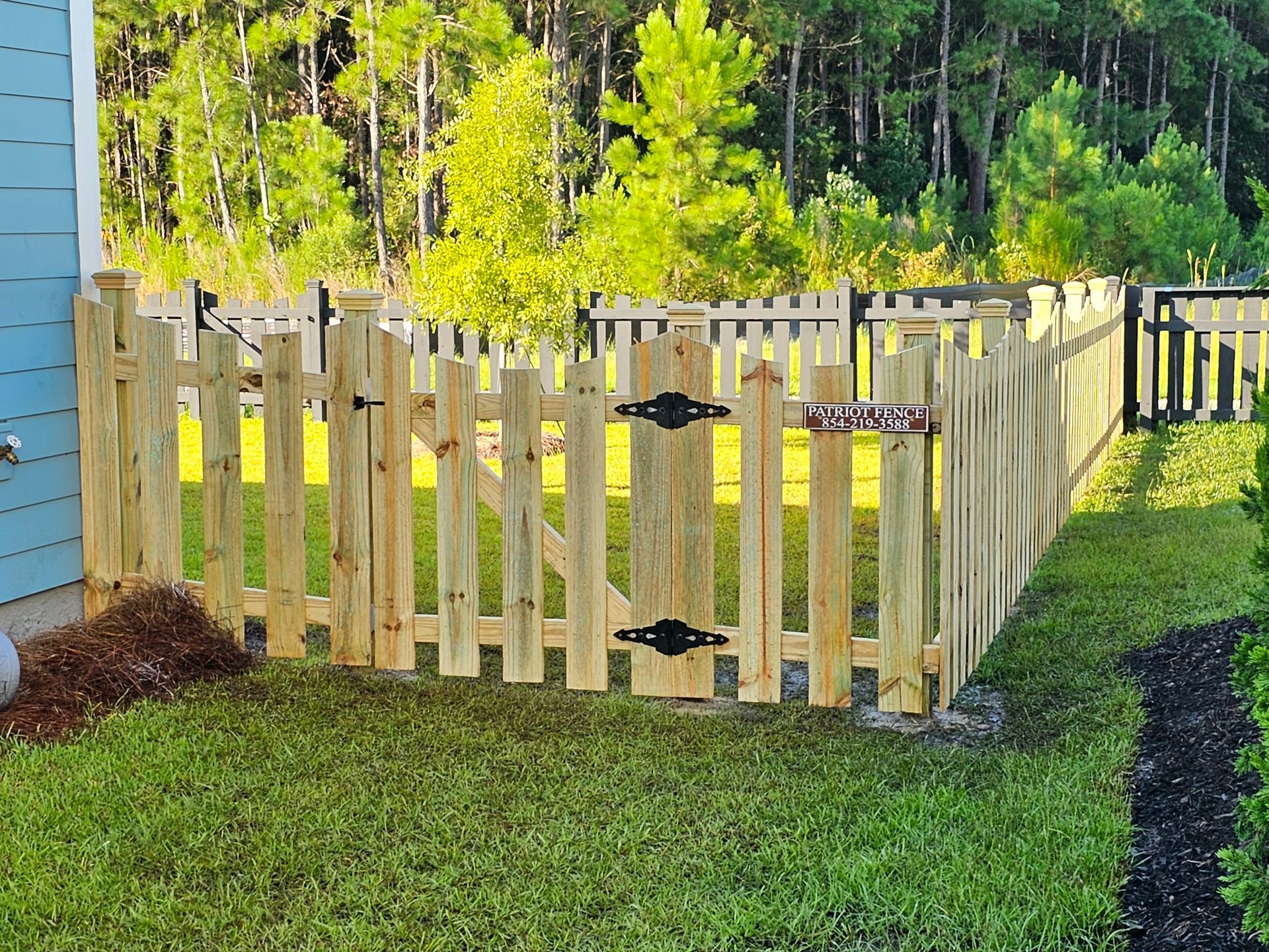 Wooden picket fence with gate in a grassy yard, surrounded by trees.