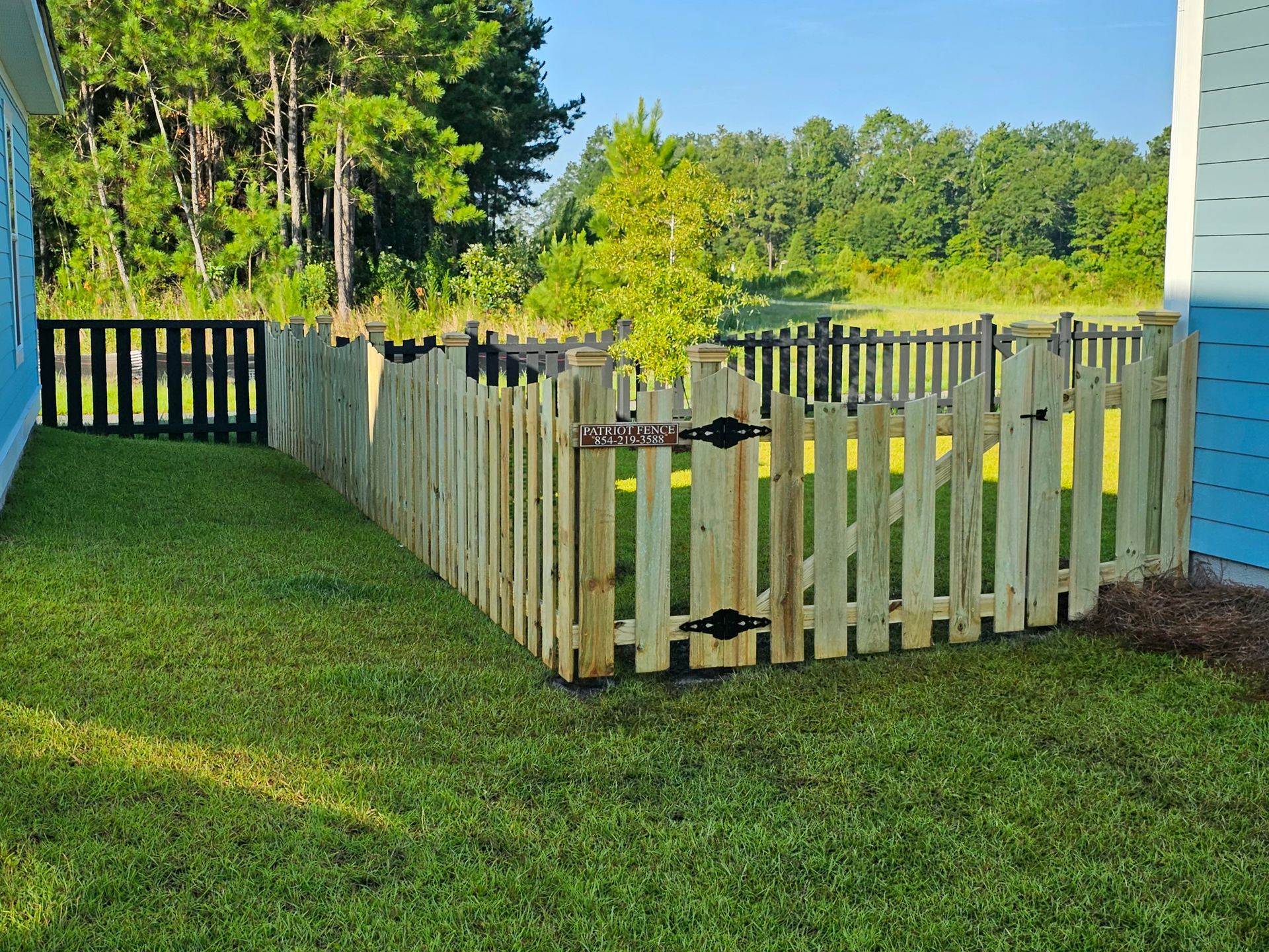 A wooden picket fence encloses a green lawn, with a black fence section and trees in the background.