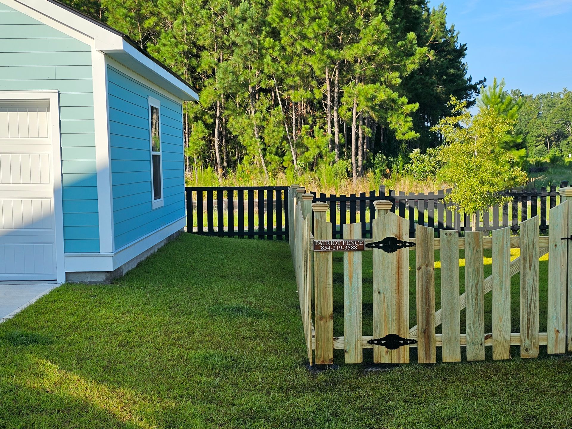 Blue house with a picket fence and a gate, overlooking a grassy yard and trees.