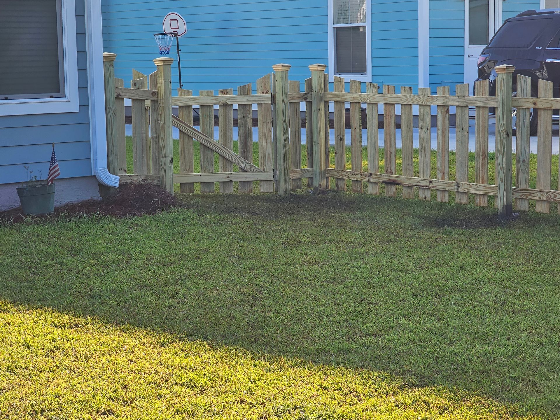 A light-colored wooden picket fence encloses a patch of grass in front of a blue house.