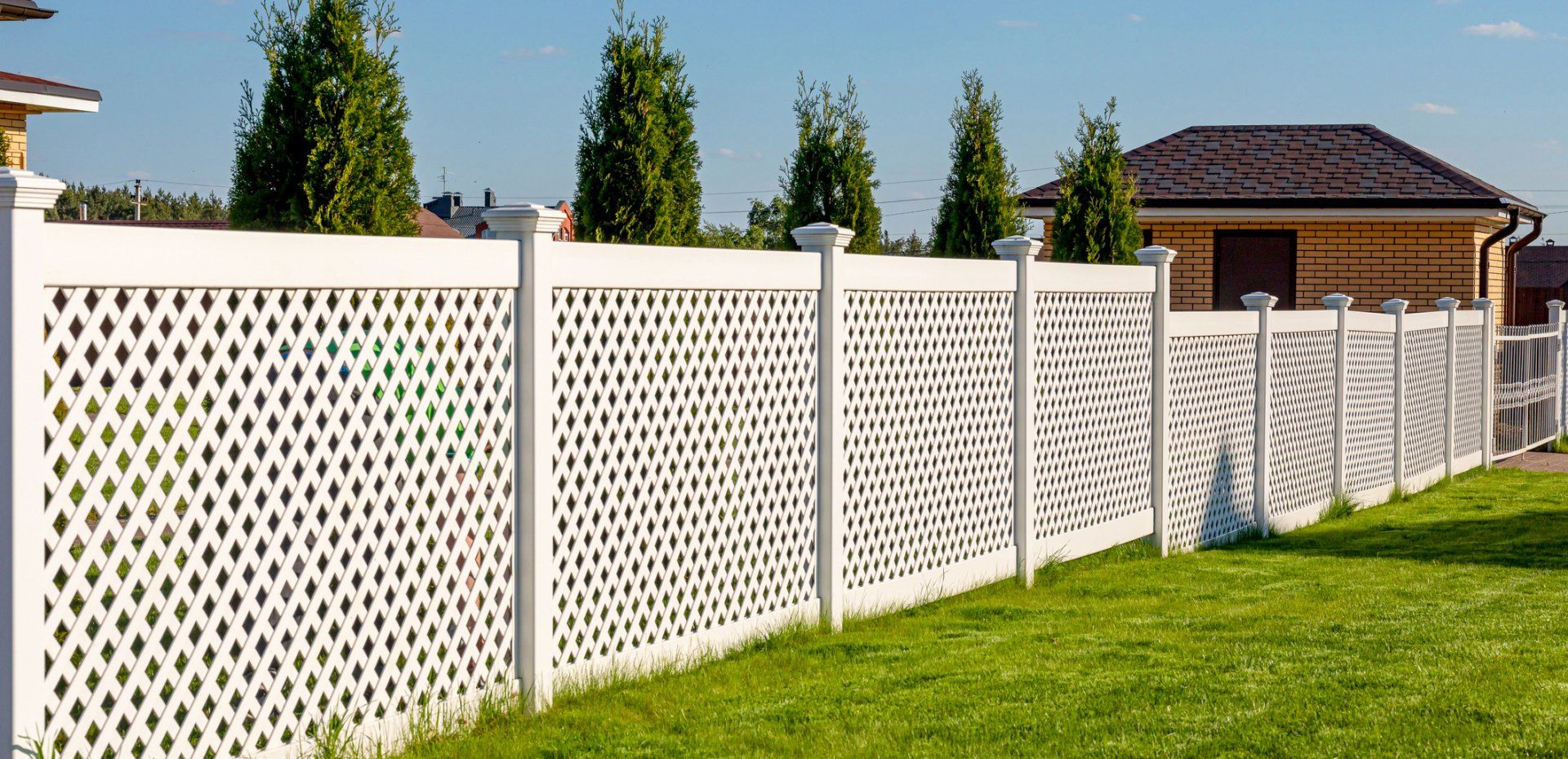 White decorative fence in a green yard with small trees and a house in the background on a sunny day.