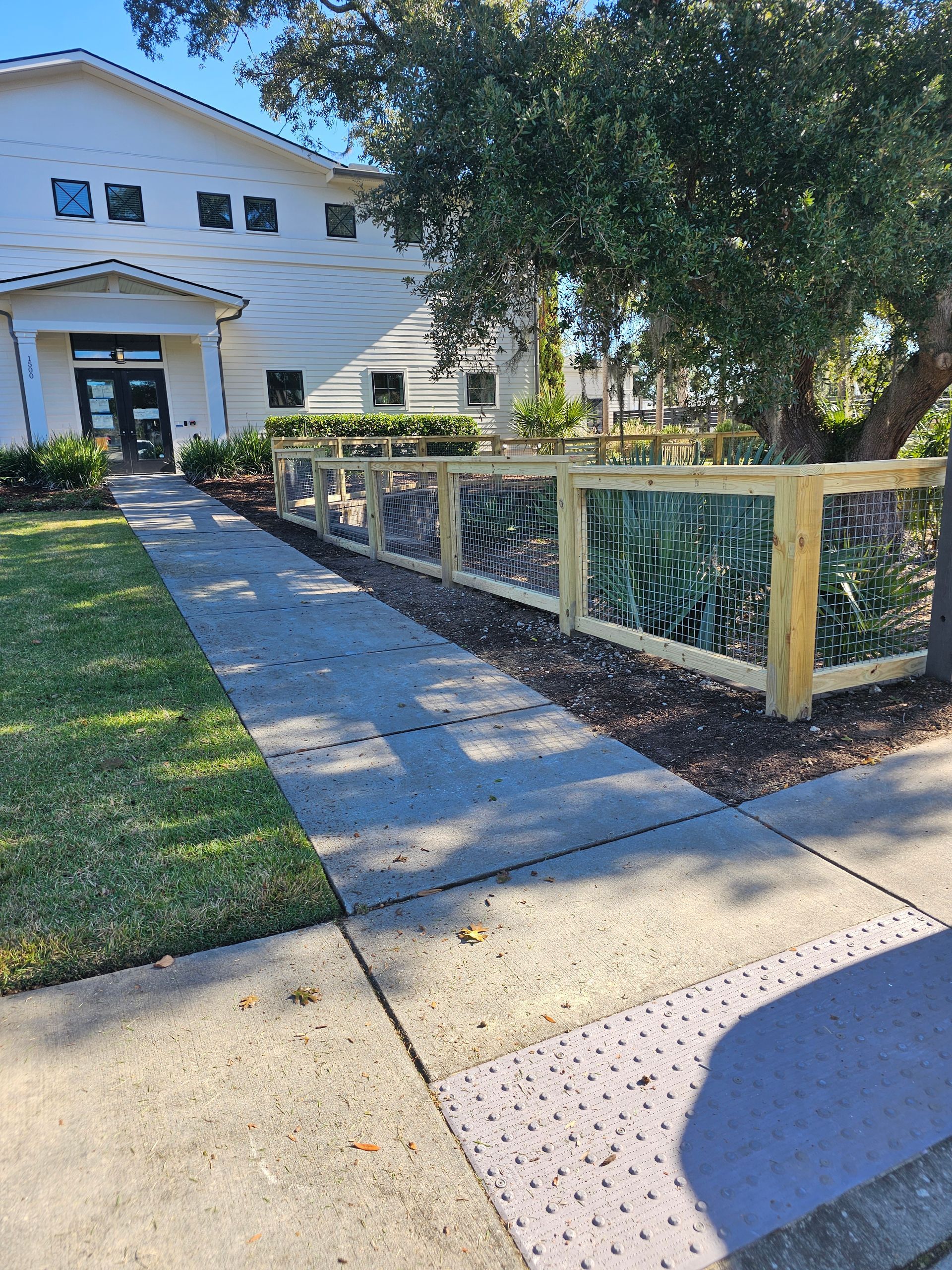 Sidewalk leading to building entrance, with a fence separating it from a planted area.