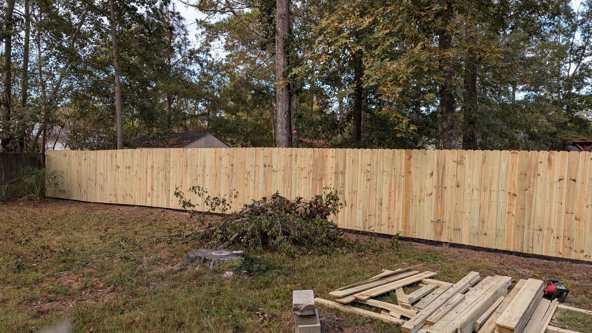 A wooden fence constructed in a yard with leaves, trees, and construction materials visible.