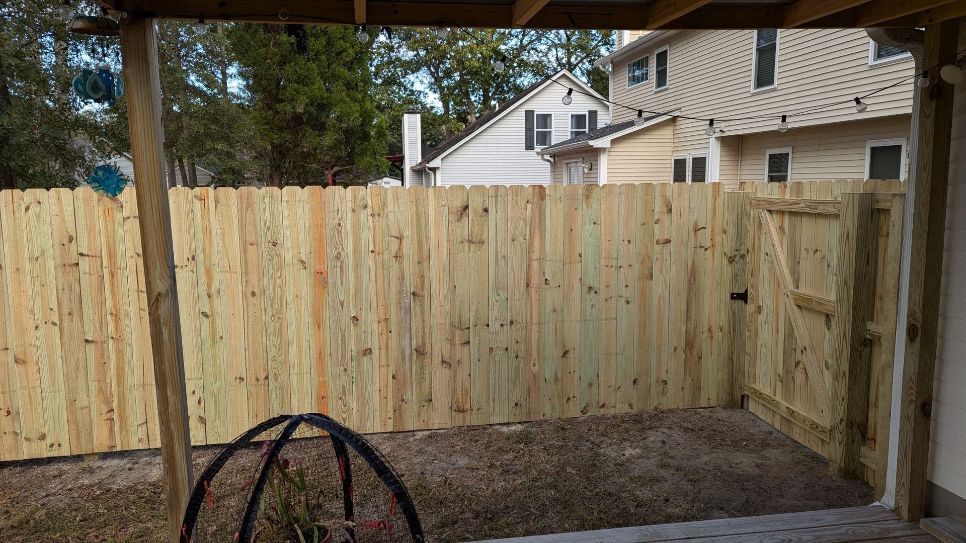 Wooden fence with gate, gravel ground, under a porch roof, houses in the background.