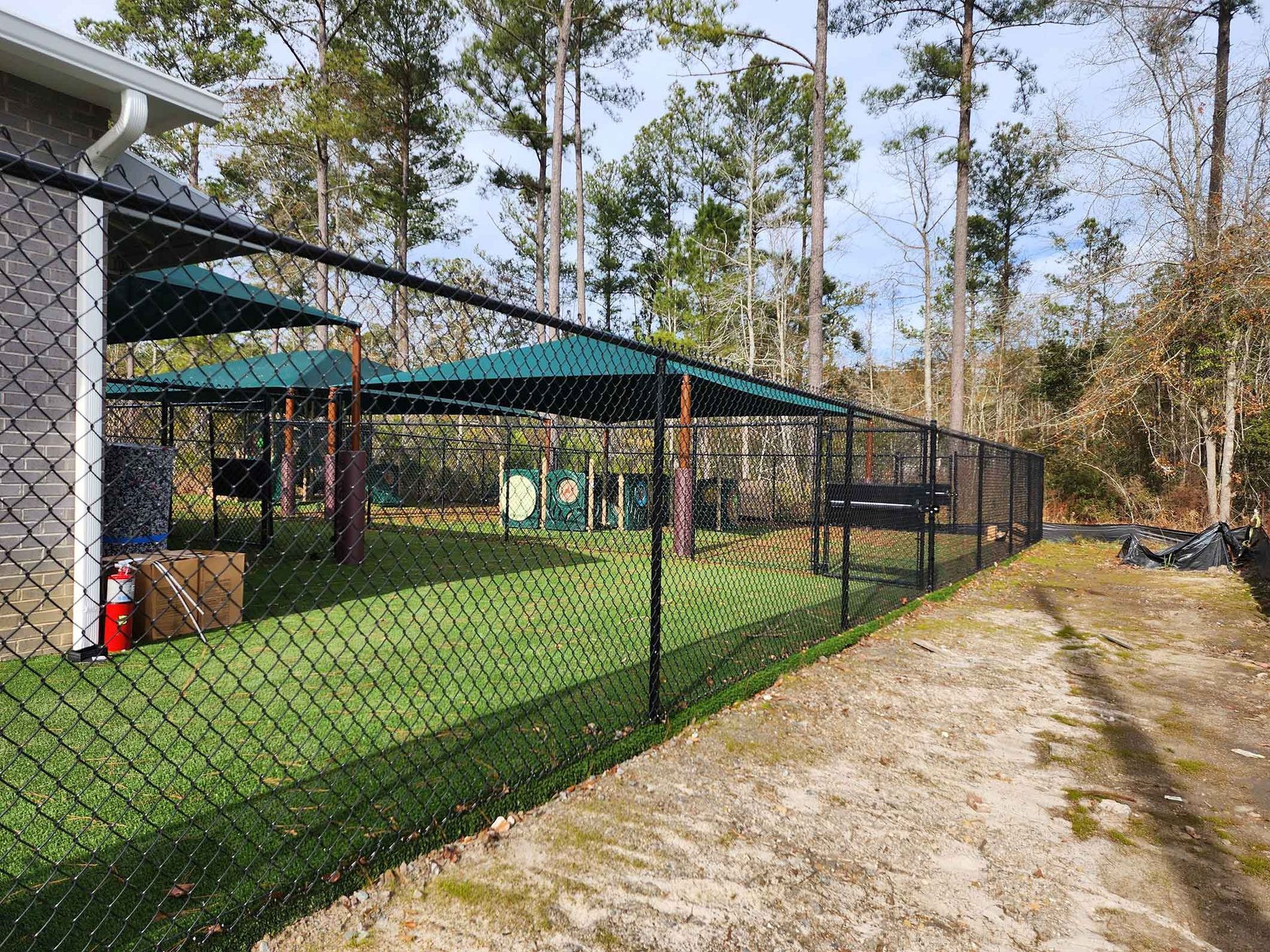 Black chain-link fence surrounds a green-roofed dog park. Green grass, trees, and a dirt path are visible.