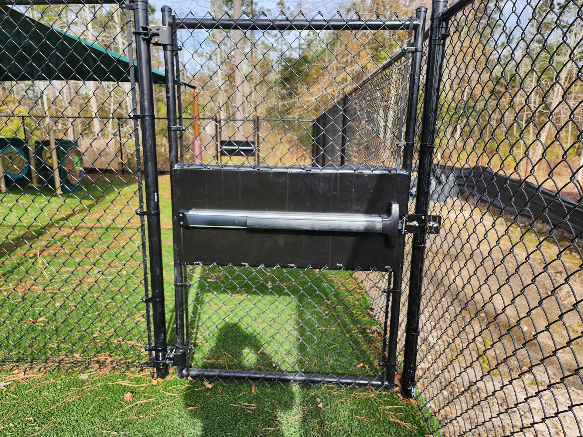 Black chain-link fence gate with a horizontal push bar in a grassy area, leading to trees in the distance.