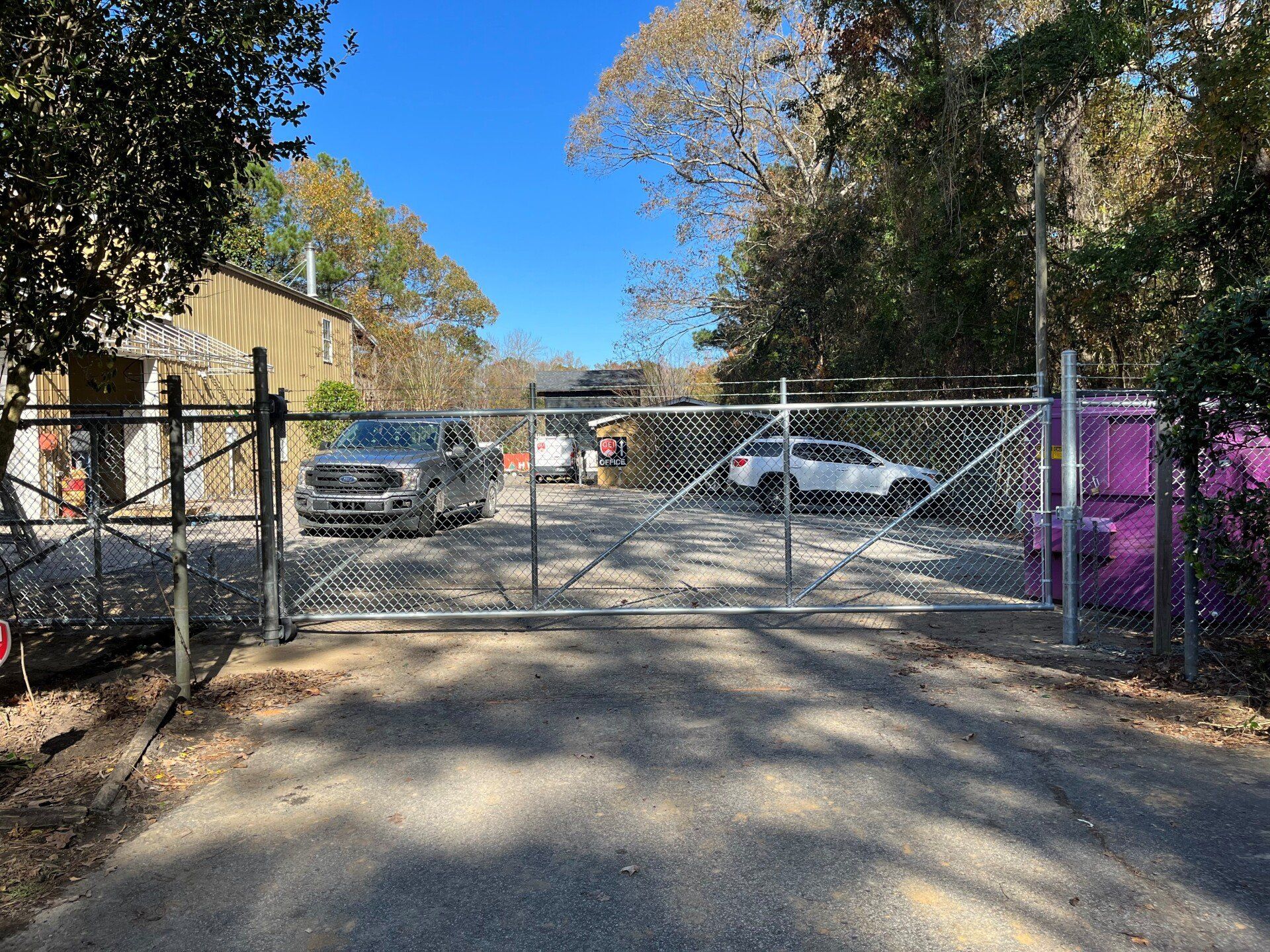 Chain link gate open, leading to a driveway with two vehicles parked in front of a building, surrounded by trees.