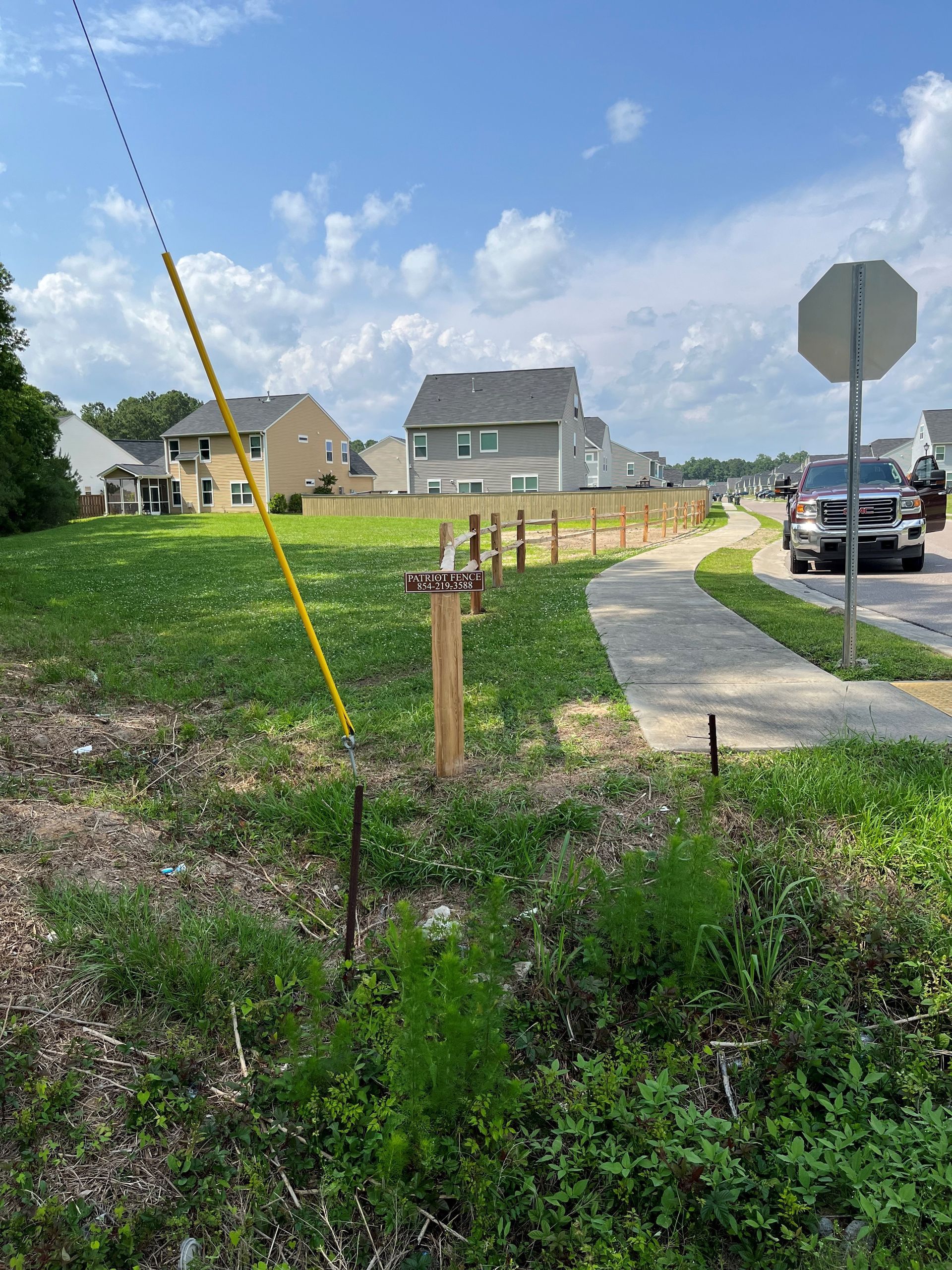 Grassy lot with sidewalk, wooden fence, houses, utility pole, stop sign, and cars on road under a blue sky.
