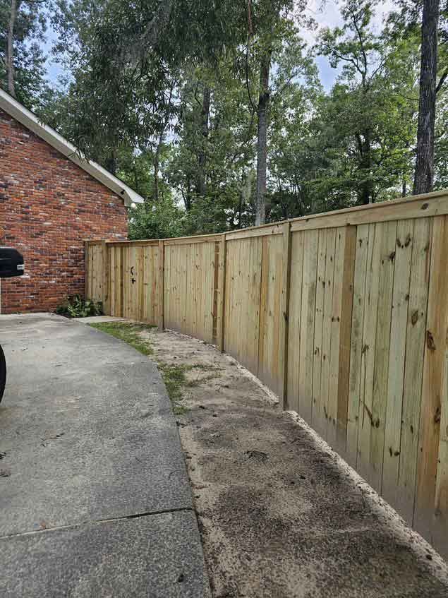 Wooden fence along a concrete driveway next to a brick building and trees.