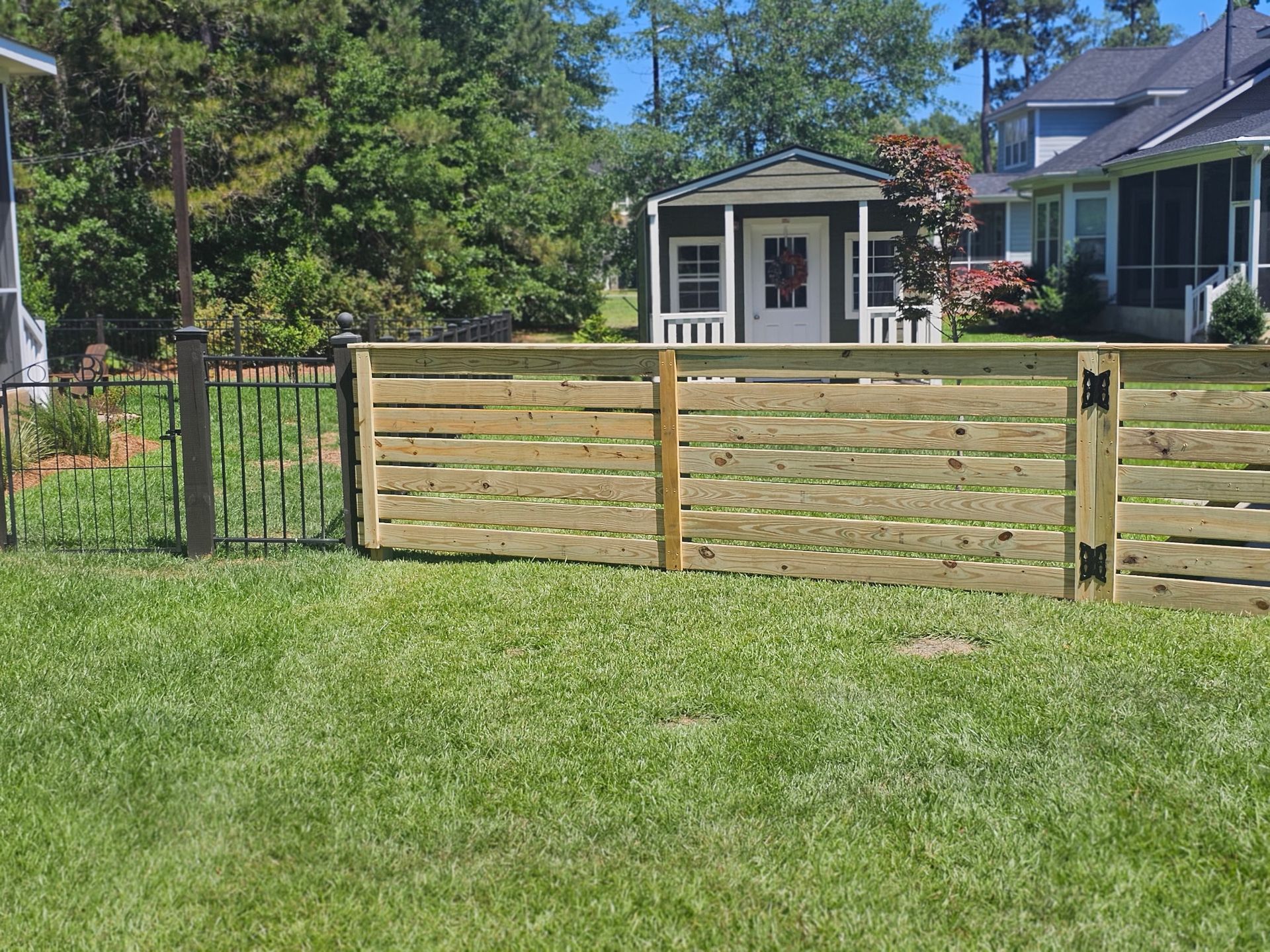 Wooden fence on grassy lawn, with a small shed and houses in the background on a sunny day.