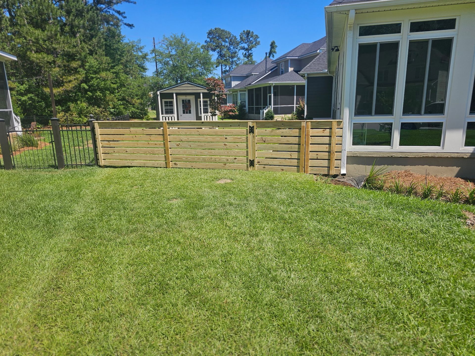 Wooden fence in a grassy yard, beside a house with large windows, on a sunny day.
