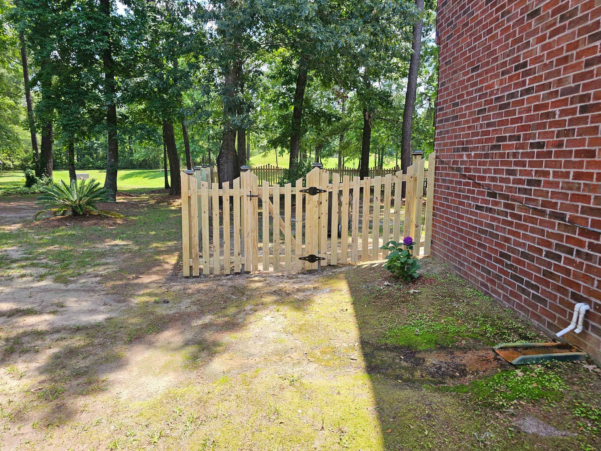 Wooden picket fence with a gate next to a red brick building, yard in background.