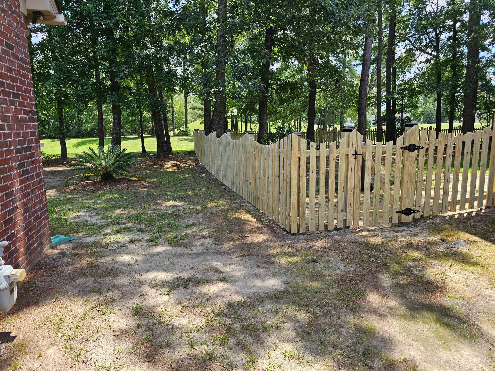 A new wooden fence surrounds a backyard with a brick wall, trees, and grass.
