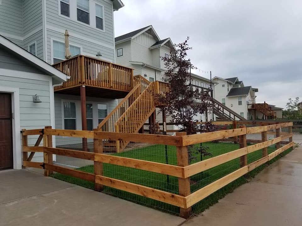 Wooden fence encloses a green lawn between multi-story buildings with wooden decks and stairs under an overcast sky.
