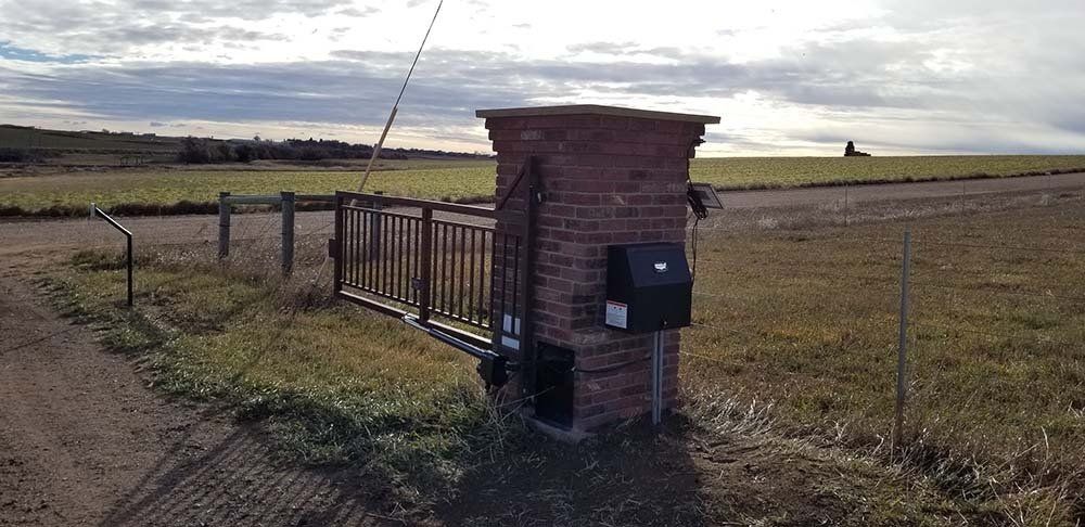 Brick pillar with a black mailbox and automatic gate on a dirt road, with grassy field and cloudy sky in the background.