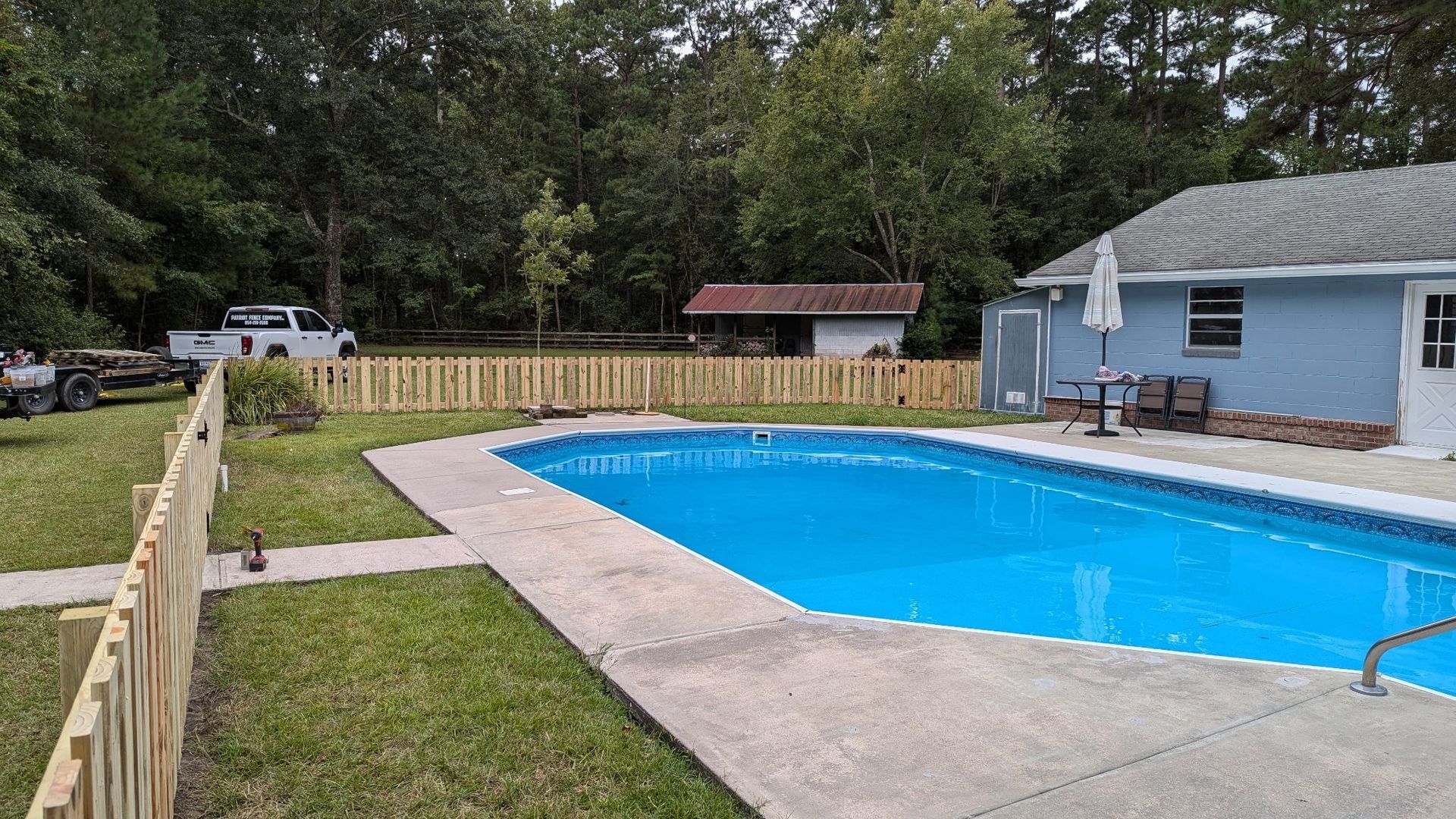 Backyard with swimming pool, fence, and light blue house. Trees in the background, grass lawn.