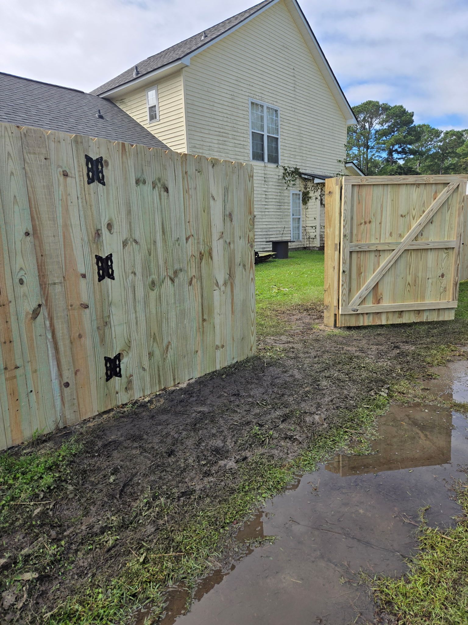 Wooden fence with gate, muddy ground. House in the background. Cloudy sky.