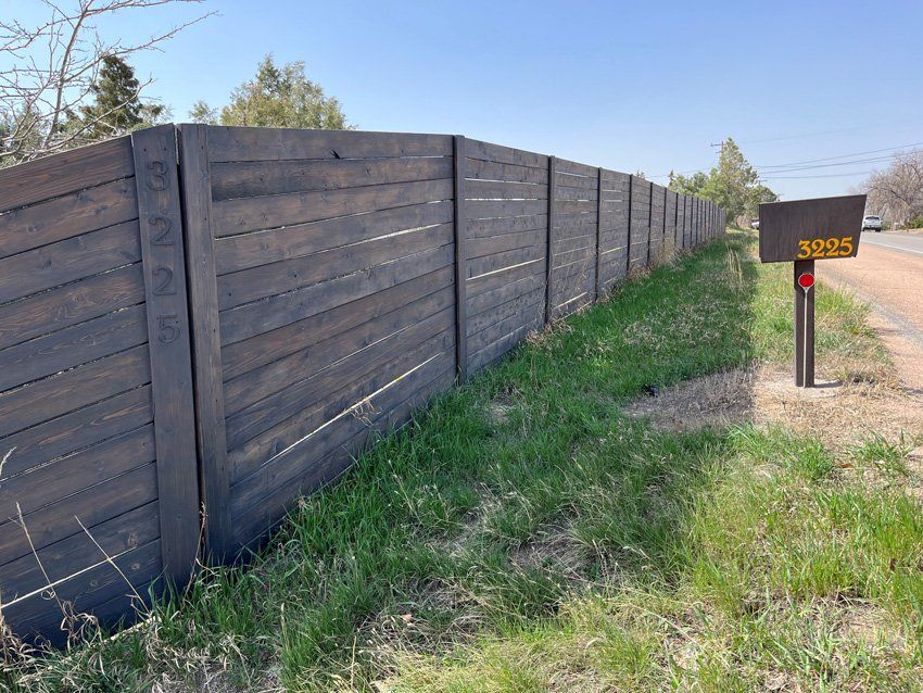 Dark wooden fence with house number 3226 on post, next to a grassy roadside.