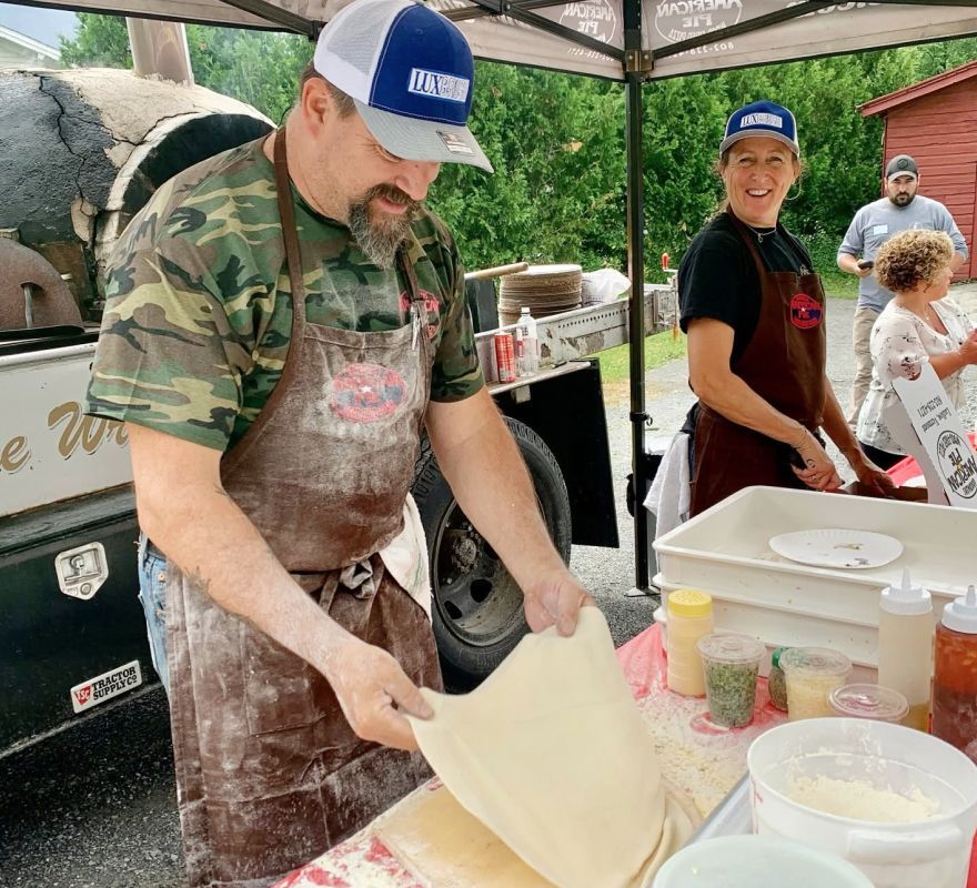 Chef stretching dough at an outdoor food stall while another volunteer smiles nearby.
